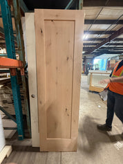 Knotty alder 1 panel Shaker door standing upright in the warehouse, blank with no handle hole cut into it, knots visible across the raw unfinished face.